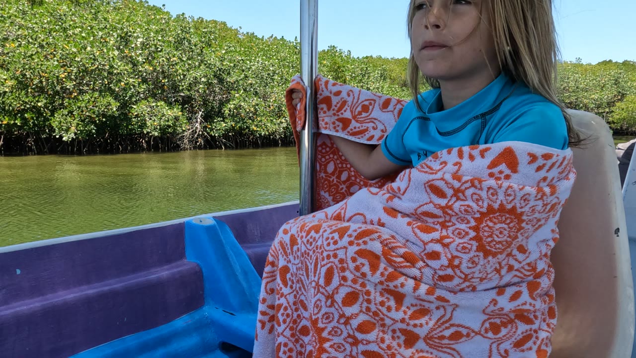 At 0:02, the girl is framed at the right edge of the boat, partly turned away, with orange blanket patterns, green water, and dense mangroves visible in bright sun.