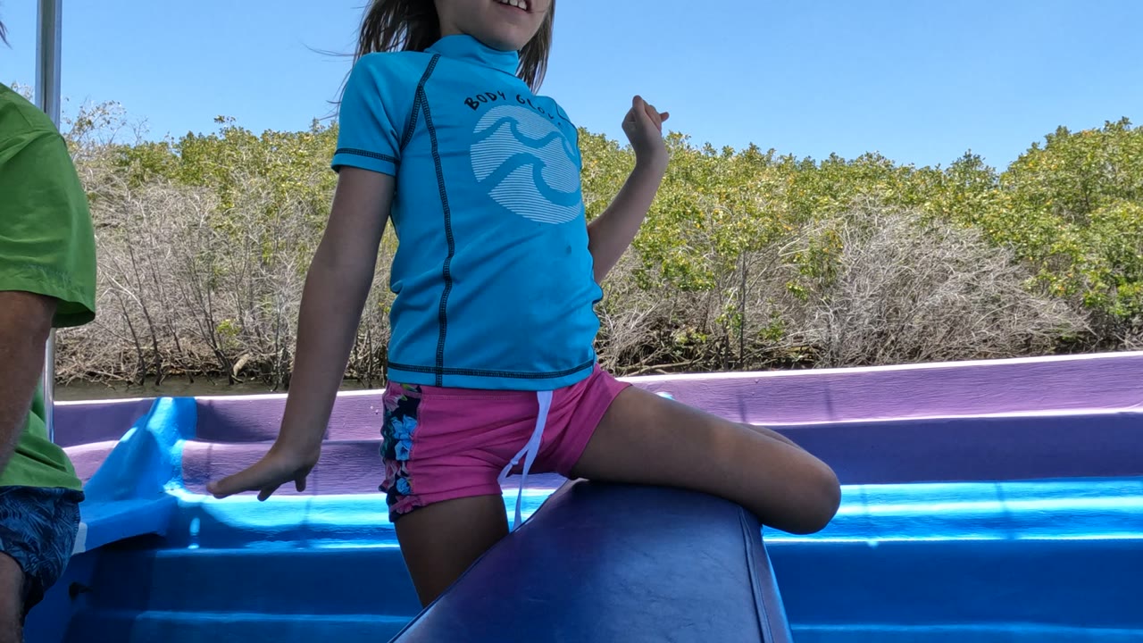 A child in a blue shirt and pink shorts stands with one knee raised on a bright blue boat in Baja California, Mexico, with another person partly visible and low trees behind the water.