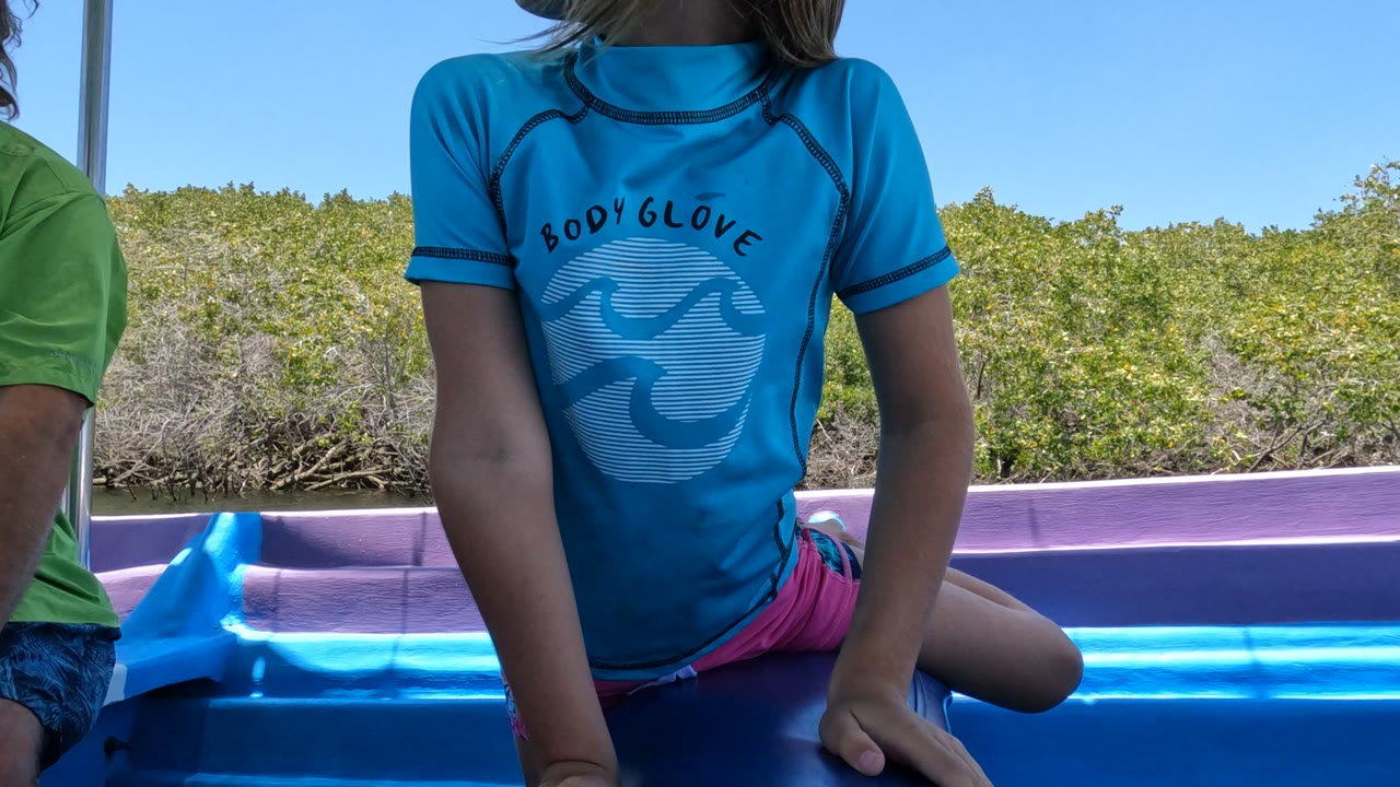 The child sits sideways on the blue and purple boat in Baja California, Mexico, with one leg bent, a cropped person nearby, and sunlit shoreline vegetation beyond the water.
