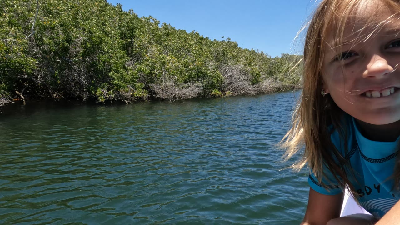 A child in a blue shirt smiles close to camera from a white boat on a river in Mulege, Mexico, with dense green trees and clear blue sky behind.