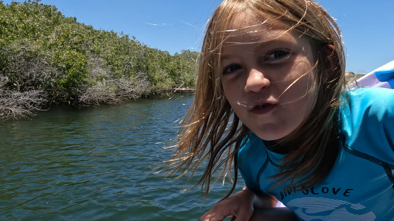 At 0:00 in Baja California, Mexico, a child in a blue shirt leans close to the camera from a small boat with rippled river water and dense trees behind them.