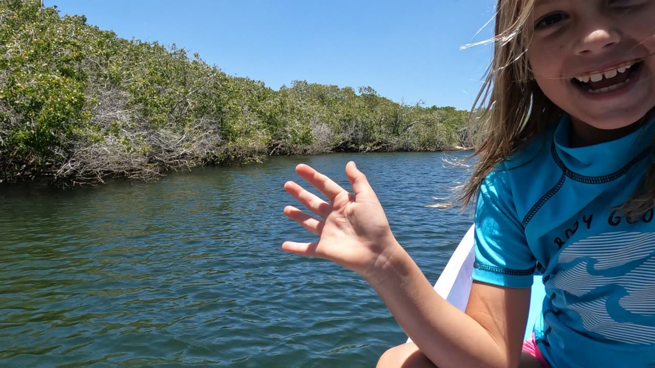 Smiling child in a blue shirt raising one hand beside river water and green trees.