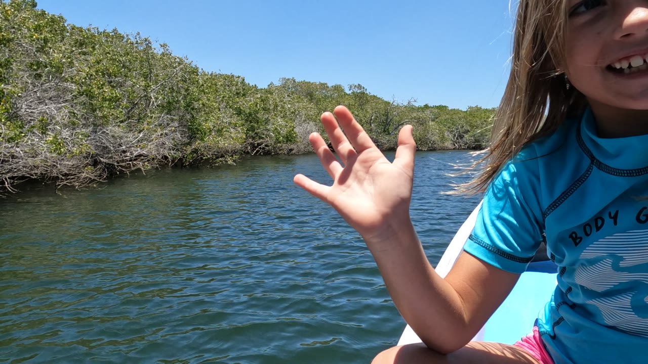 At 0:02 in Baja California, Mexico, the child is partly cropped on the right side of the frame while an open hand is held up against water, trees, and blue sky.