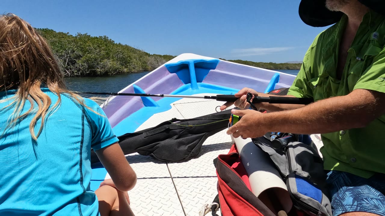 In Baja California, Mexico, a girl in a blue shirt sits at the front of a small boat while a man in a green shirt holds a fishing rod over boat gear near calm water and low shoreline trees.