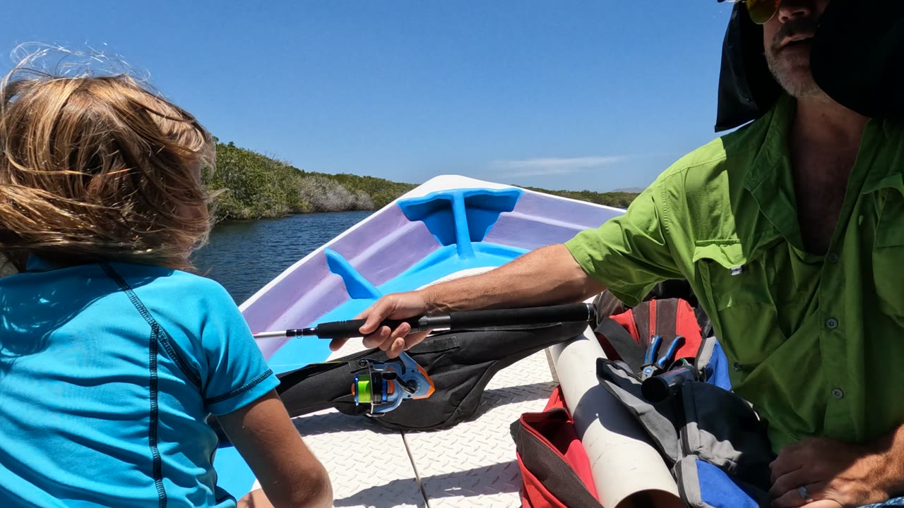 In Baja California, Mexico, the girl turns her back toward the camera as the man extends the rod across the boat, with the bright bow, black gear, water, and green shoreline behind them.