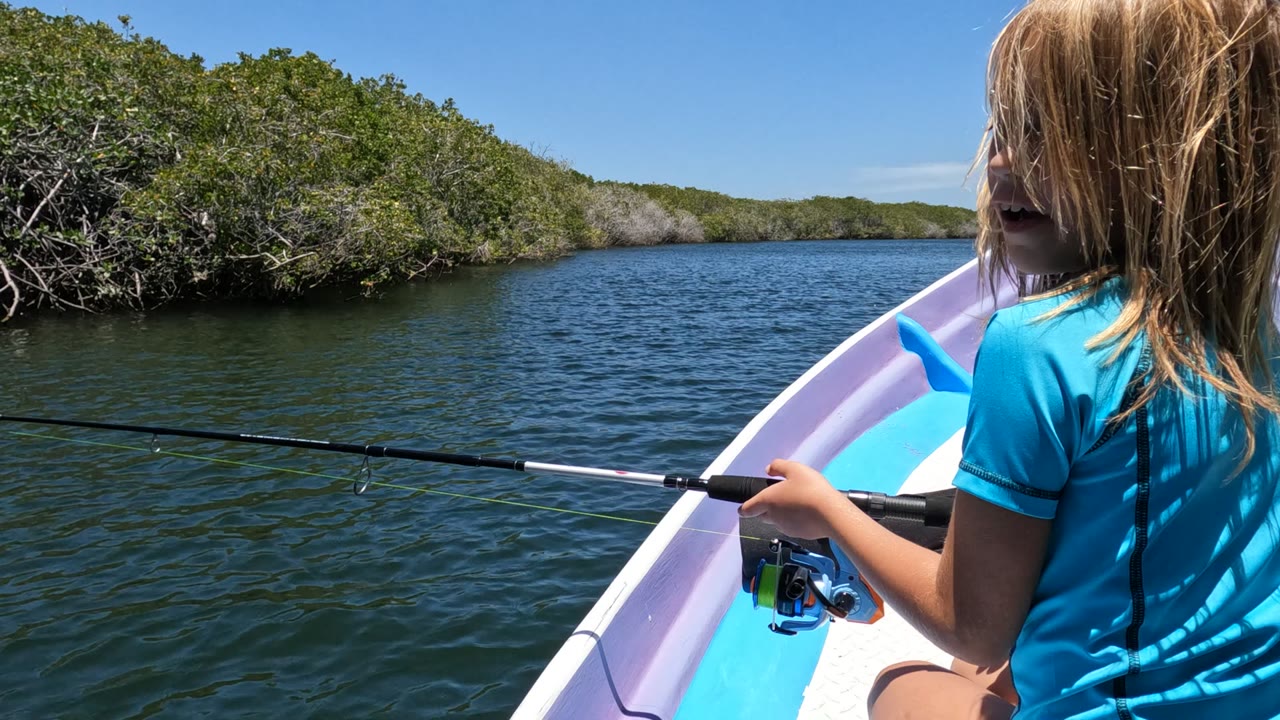 The girl remains partly visible on the right side of the boat in Baja California, Mexico, with the fishing rod crossing open water and mangroves filling the far bank.