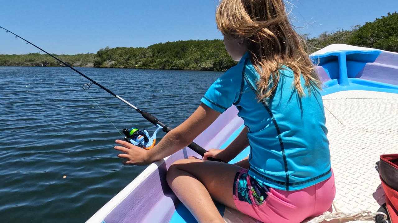 At 0:00, a girl in a blue shirt and pink shorts sits near the side of a small boat in Baja California, Mexico, with a fishing rod angled over rippled water and green shoreline beyond.