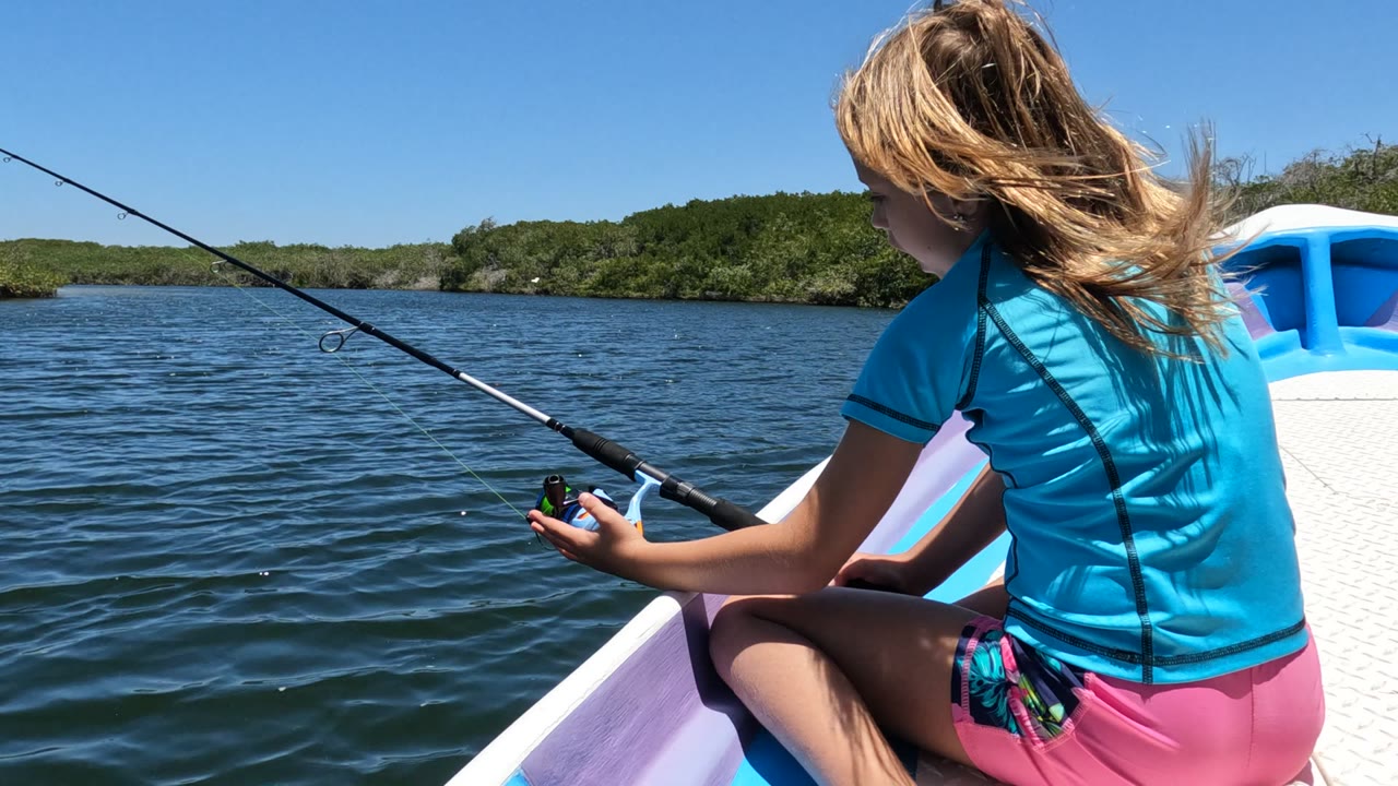 At 0:02, the girl sits on the right edge of the frame, her fishing rod stretching diagonally across open water toward a low green shore in Baja California, Mexico.