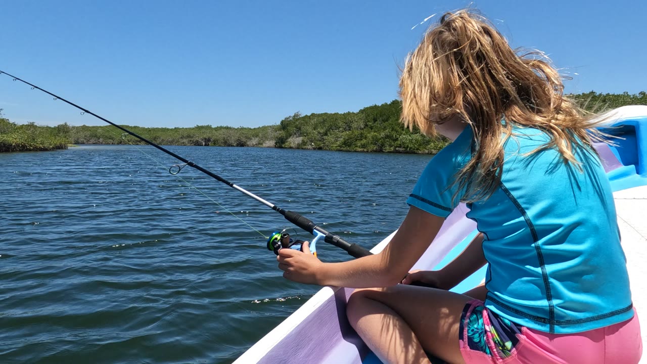 At the start of the clip, a girl in a blue shirt sits in a small boat in Baja California, Mexico, holding a fishing rod over rippled water with a green shoreline ahead.