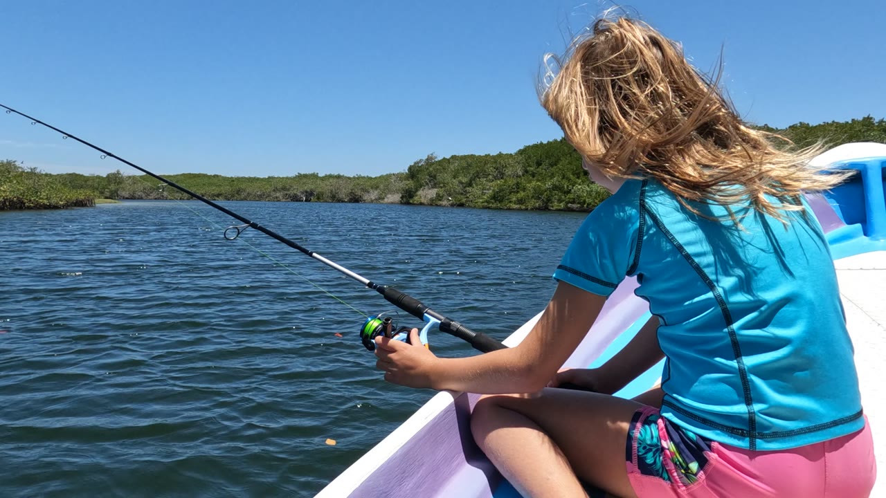 At 0:02, the girl remains seated near the boat edge with the rod extending left over the water, and the open channel and green shoreline stay level in the distance.