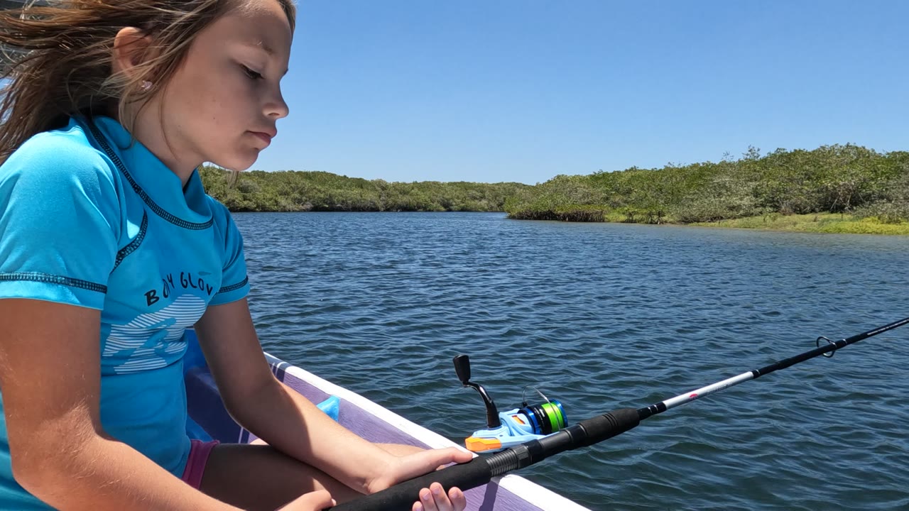 At the start of the Baja California, Mexico clip, a girl in a blue shirt sits at the left edge of the boat and holds a fishing rod over rippled water with low green shoreline behind her.