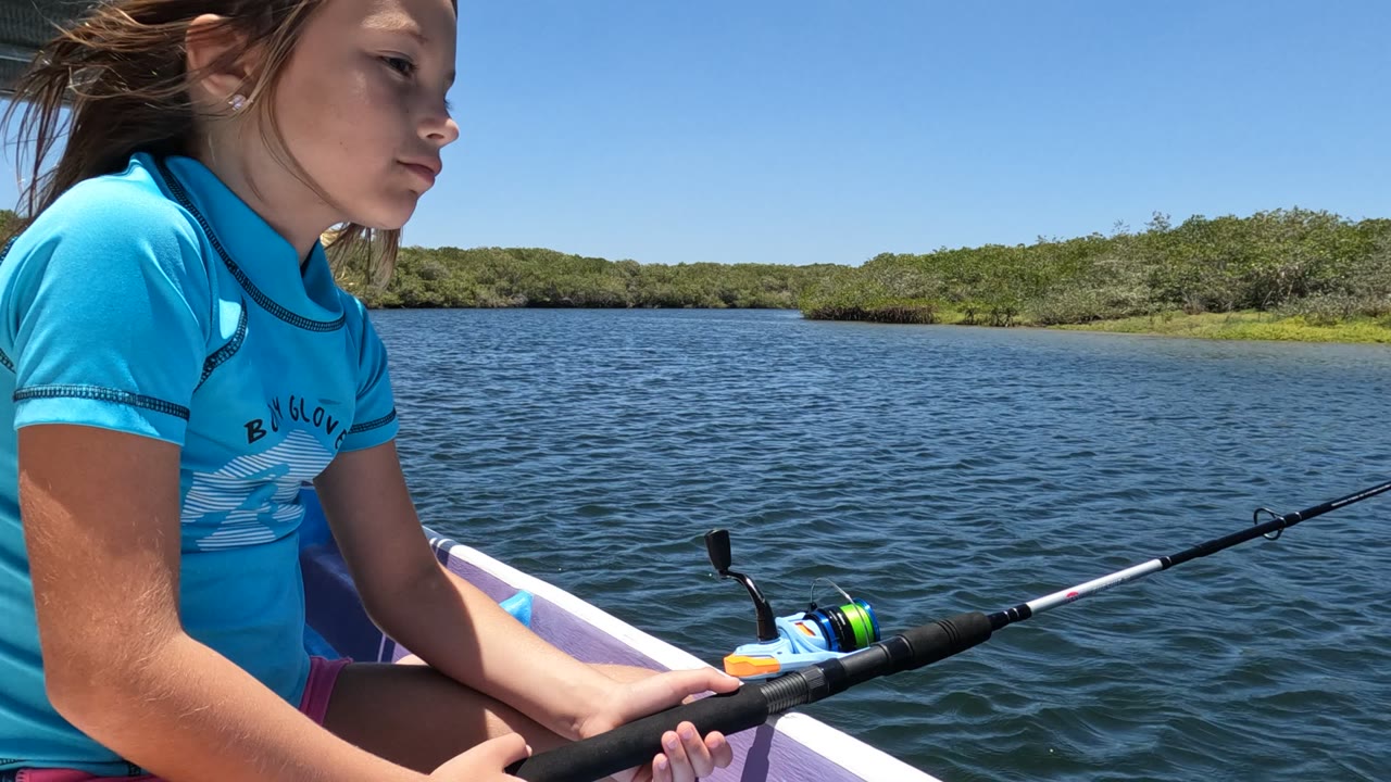 At 0:02, the girl faces right in profile with the fishing rod extending diagonally across the frame over blue water and a low green shore in Baja California, Mexico.