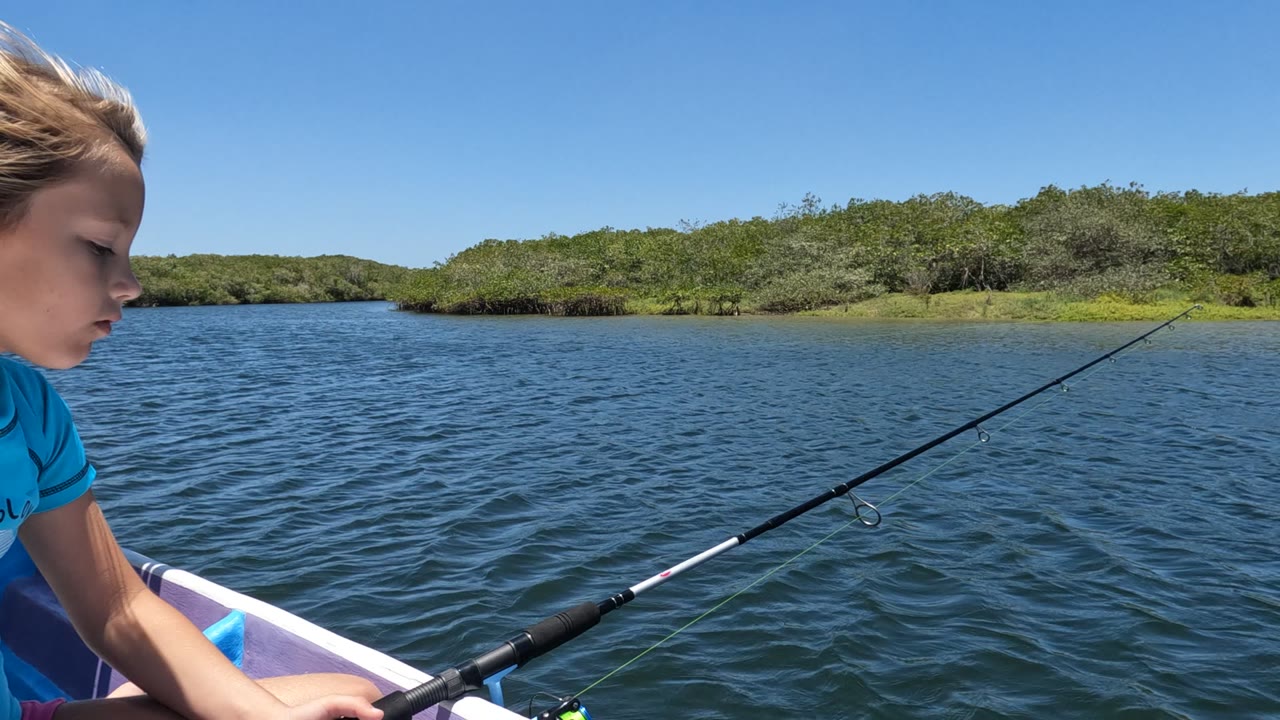 At the start of the Baja California, Mexico clip, a girl in a blue shirt is cropped at the left edge while a fishing rod points across rippled water toward a low green shore.