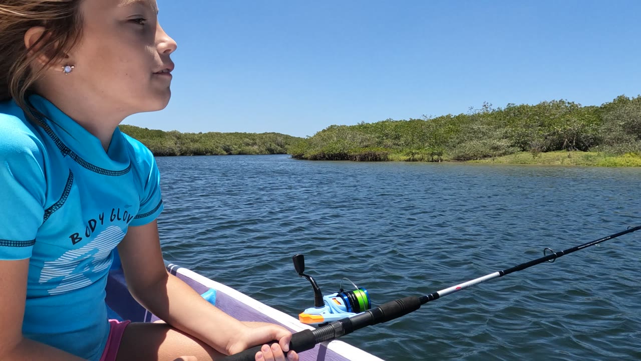 At 0:02, the girl appears in side profile with her face turned toward the water, while the fishing rod and reel sit across the foreground against bright blue water.