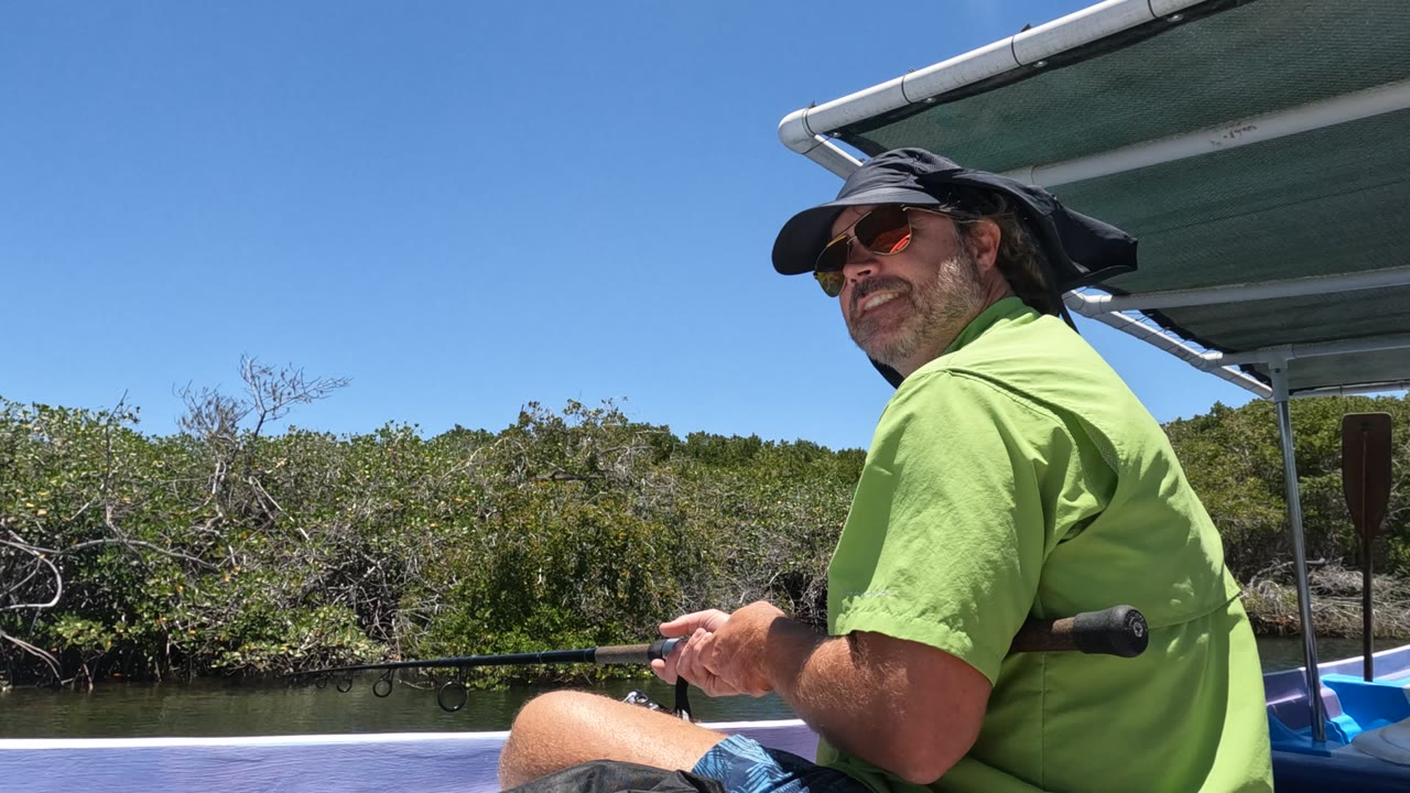 A man in a green shirt, dark hat, and sunglasses holds a fishing rod from a boat on a calm river in Baja California, Mexico, with dense trees behind him.