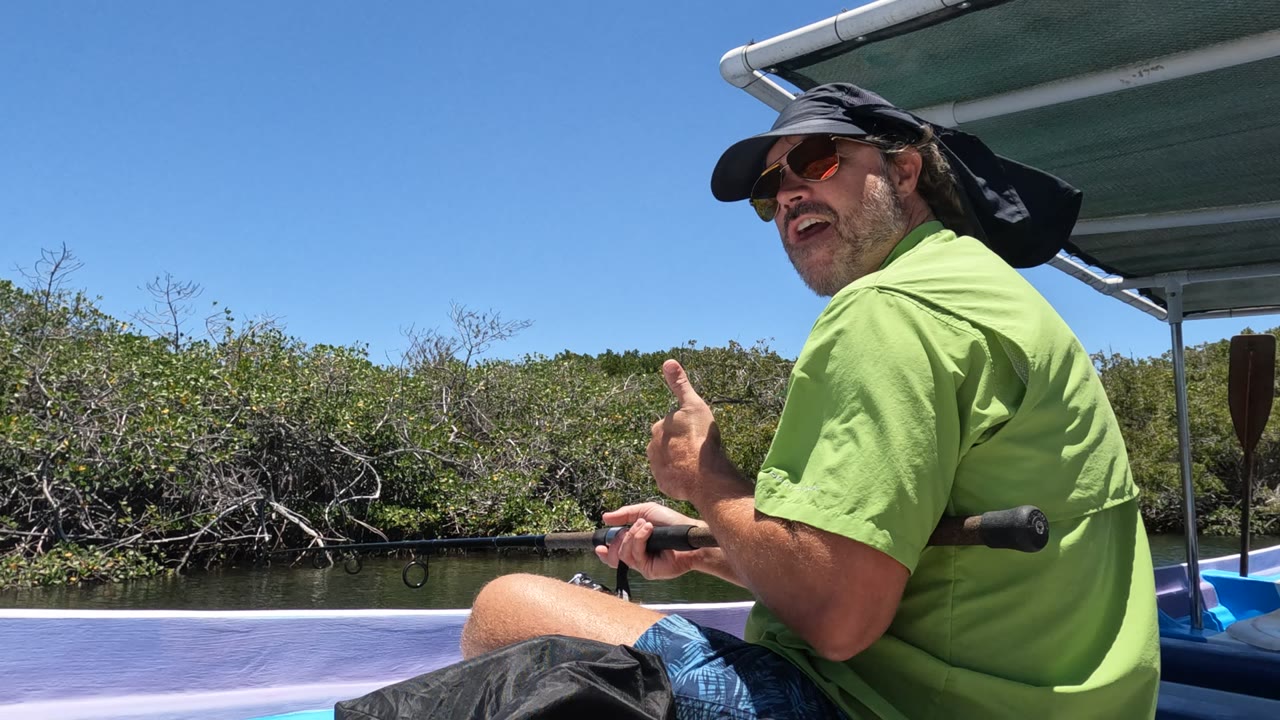 A man in sunglasses and a green shirt gives a thumbs-up while seated with a fishing rod on a boat in Baja California, Mexico, near low green riverbank vegetation.