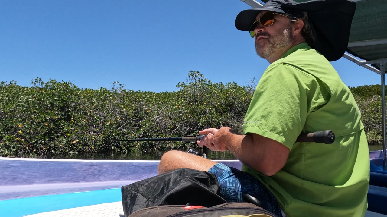A man in a green shirt, sunglasses, and a wide sun hat sits on a boat in Baja California, Mexico, holding a fishing rod with mangroves behind him.