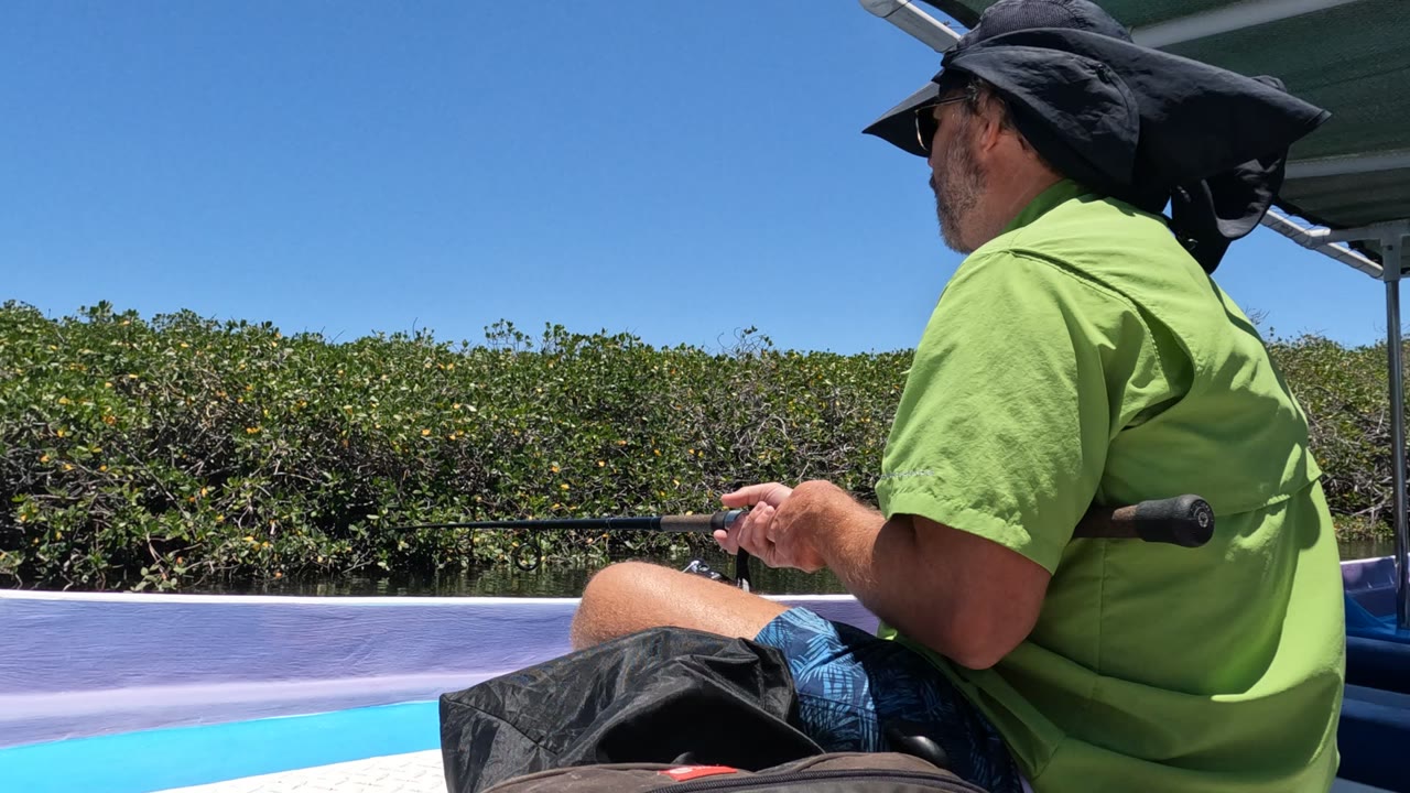 The man turns more away from camera while seated on the boat, with his green shirt, dark hat flap, fishing rod, mangroves, and clear sky visible in Baja California, Mexico.
