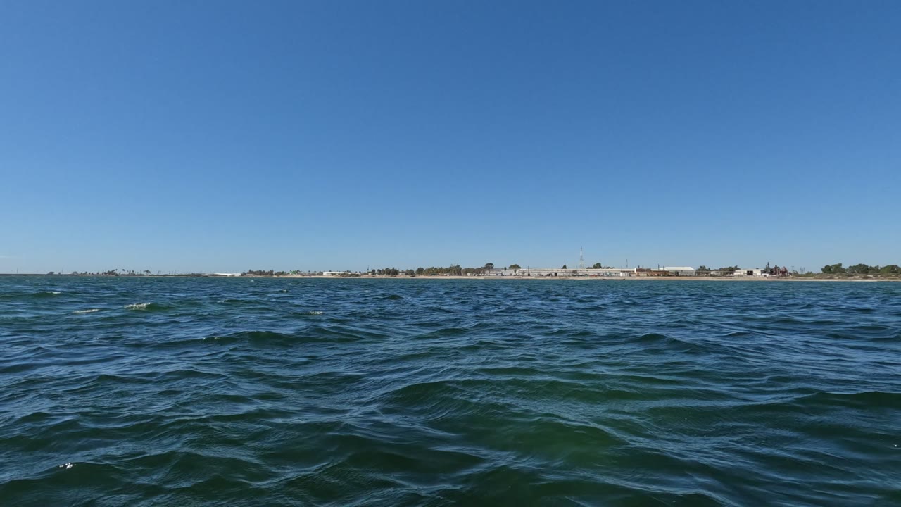Baja California ocean water fills the foreground with small blue-green waves, a thin shoreline and buildings sit on the horizon, and no visible people appear.