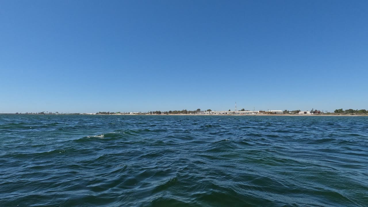 A darker swell crosses the midground of the Baja California ocean view, with low buildings visible along the far horizon and no visible people.