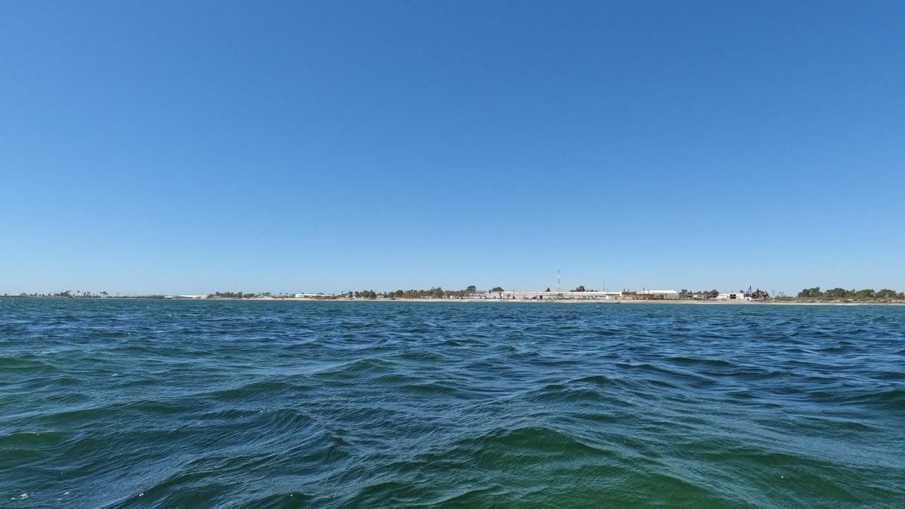 A slightly shifted wave pattern crosses the foreground water in Baja California, Mexico, while the distant shore remains thin beneath a cloudless sky, no visible people.