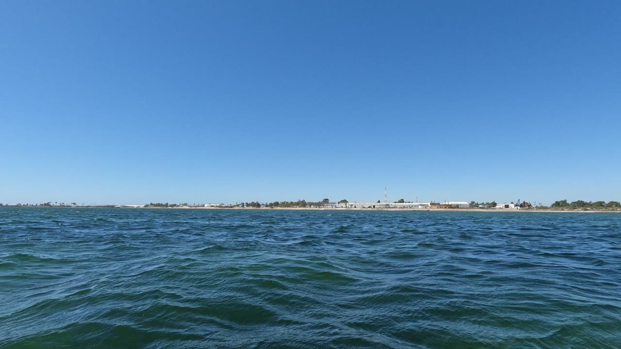 Rolling blue water sits low in the frame in Baja California, Mexico, with a long sandy shoreline, scattered low structures, and no visible people.
