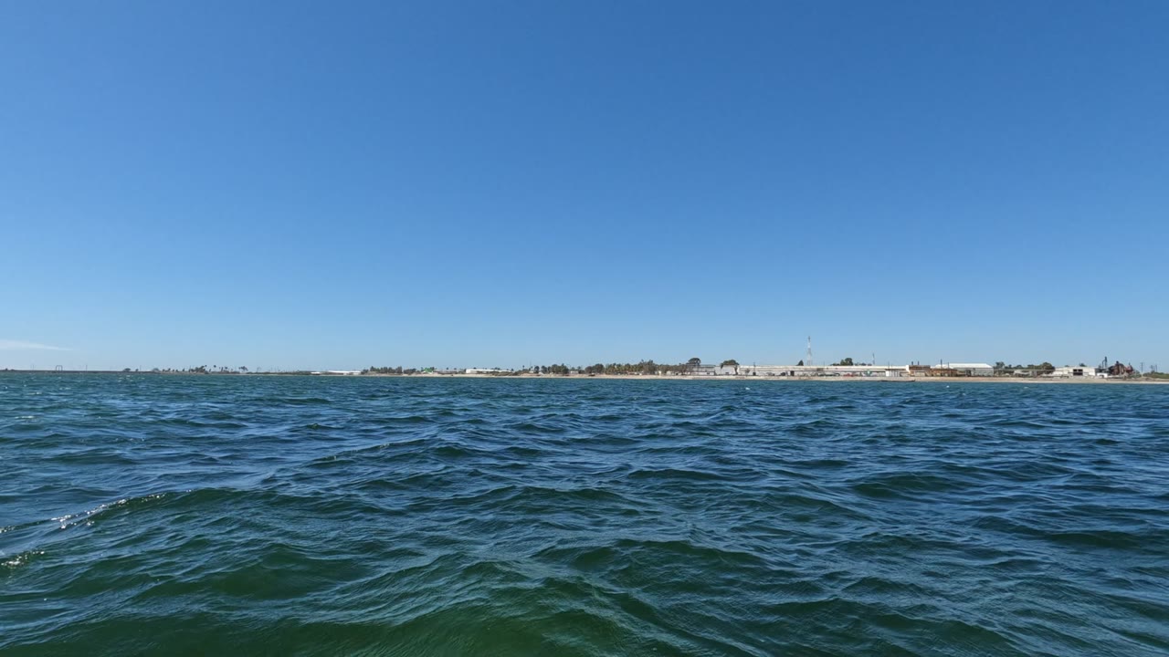 Baja California ocean water fills the foreground at the start of the clip, with a thin distant shoreline and low buildings on the horizon and no visible people.