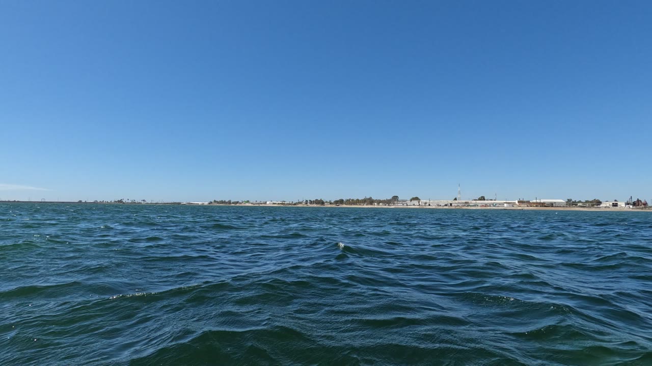 At 0:02, small ocean swells catch daylight in the foreground while the Baja California shoreline remains a narrow band of land and low buildings in the distance, with no visible people.