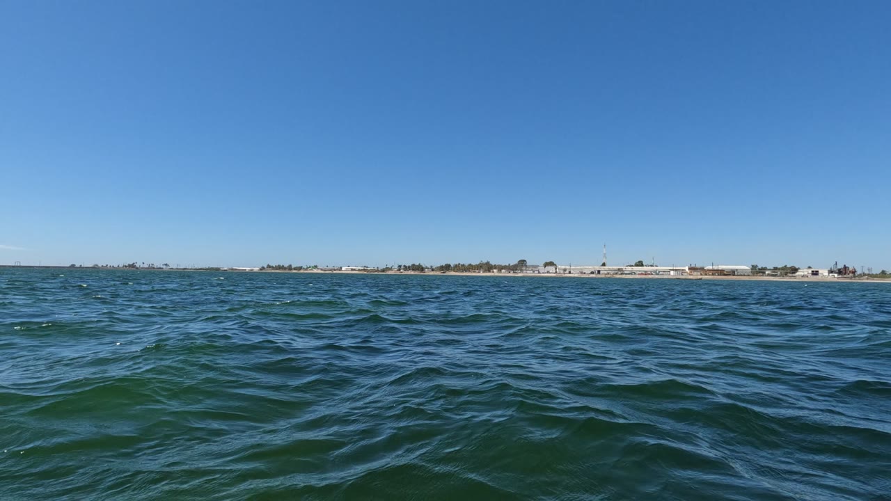 Baja California ocean water fills the foreground with small waves, a thin shoreline with low buildings crosses the horizon, and no visible people appear at the start of the clip.