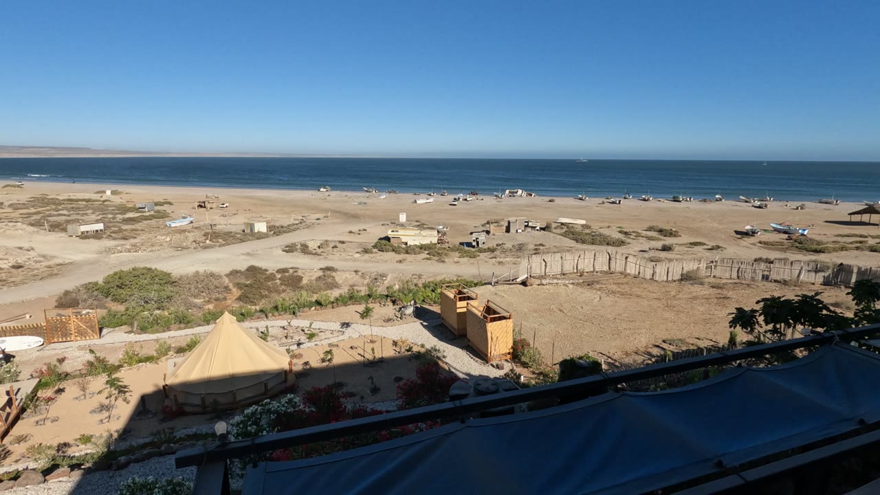 Wide elevated view of a Baja California beach camp with canvas tents, small structures, sandy ground, ocean horizon, clear blue sky, and no visible people.
