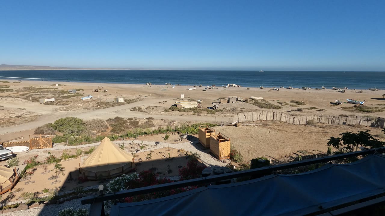 Wide Baja California beach view with tents and wooden structures in the lower sand, scattered boats near the shore, a dark blue ocean band, and no visible people.
