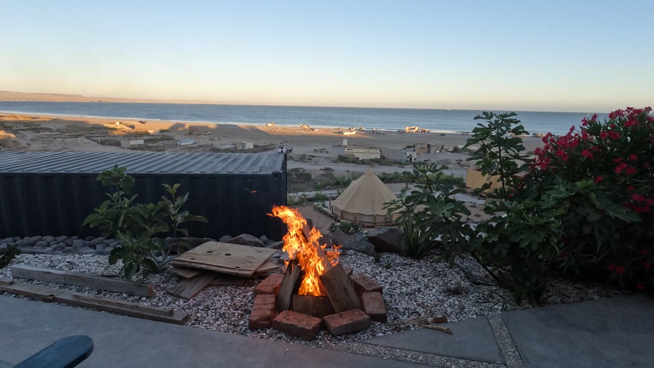A Baja California campfire burns in a brick ring on a gravel patio, with red flowers, a dark container structure, a tent, beach, ocean, and no visible people.