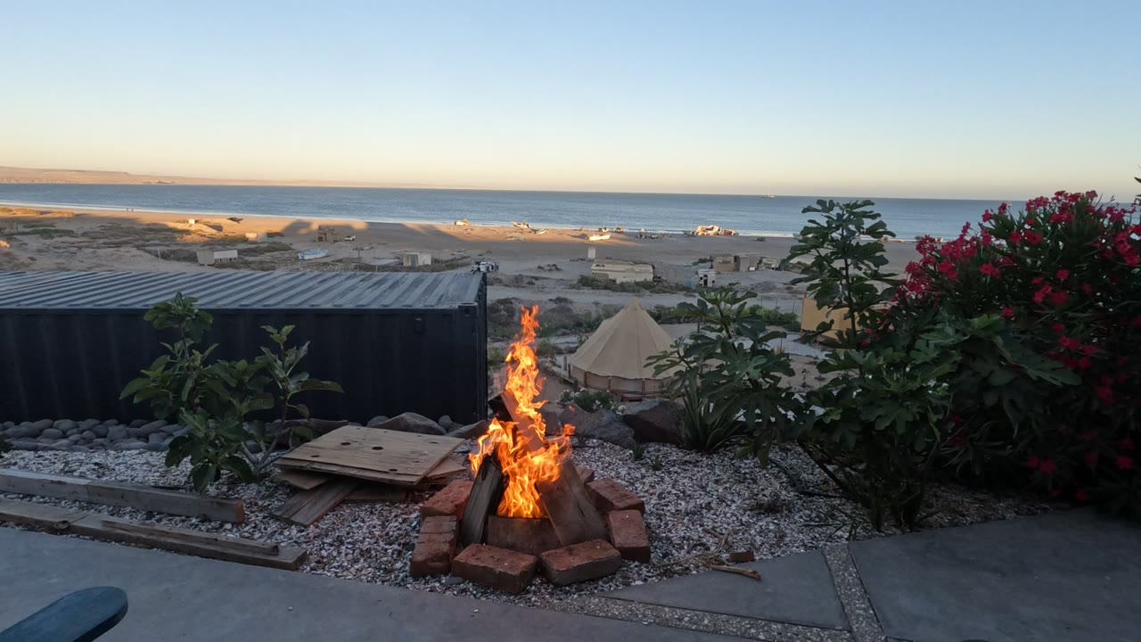 Tall flames rise from a brick-ring fire with beach and ocean in the background.