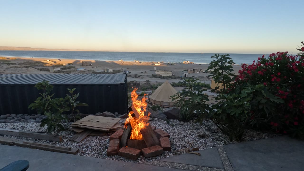 The Baja California campsite view shows the flame narrower in the brick ring, red flowers to the right, a dark container structure to the left, a tent beyond, ocean water, and no visible people.