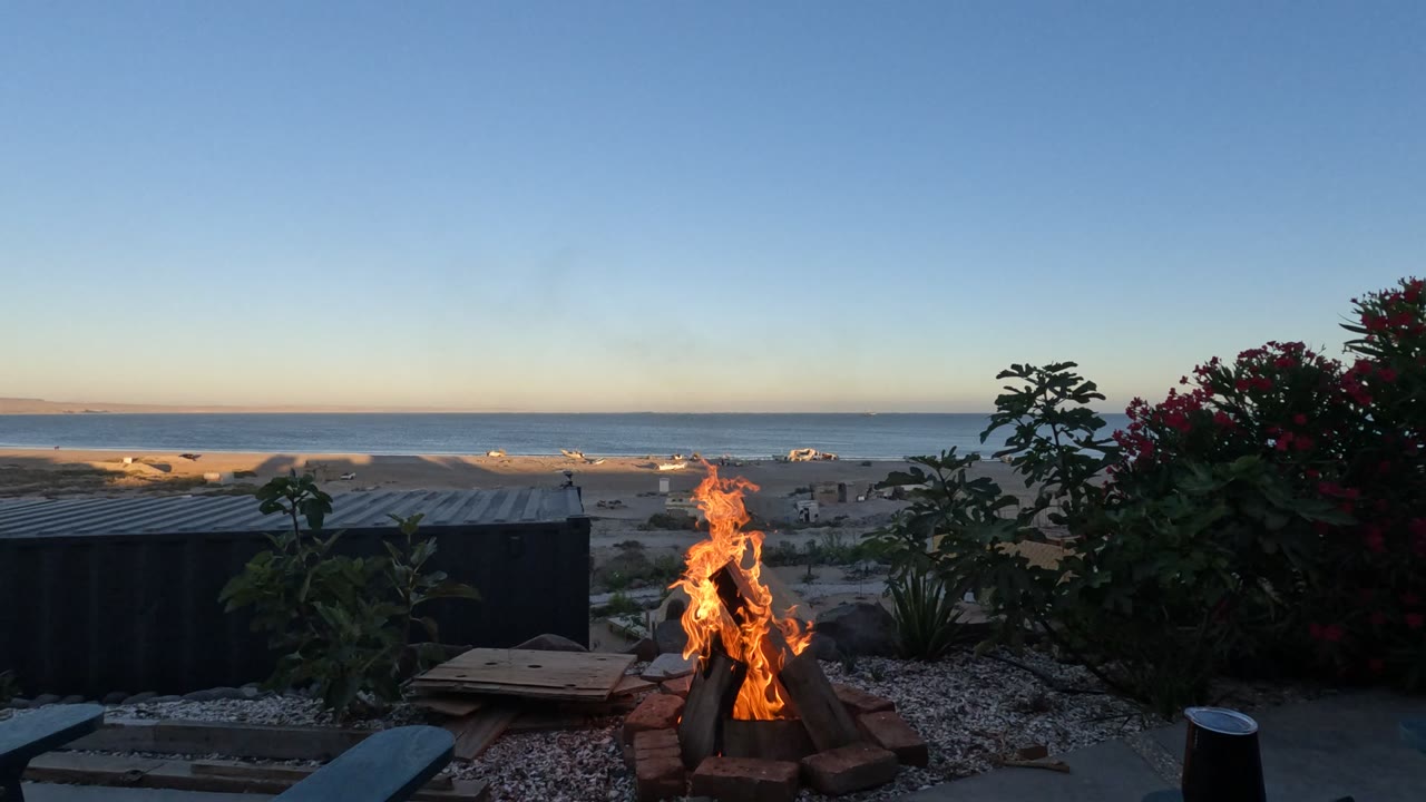 A fire pit burns beside gravel and patio seating in Baja California, Mexico, with red flowers, a low beach structure, ocean horizon, pale dusk sky, and no visible people.