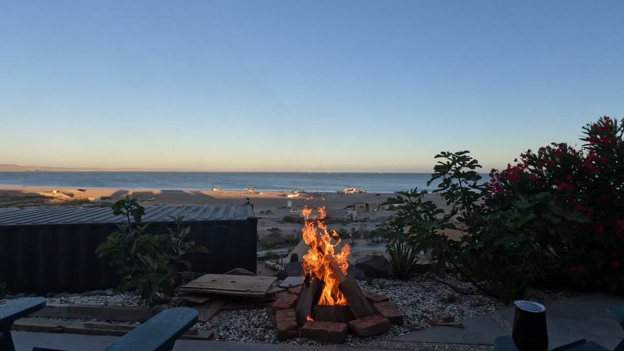 A beach fire pit burns lower in Baja California, Mexico, while a thin column of smoke drifts upward above the sand, ocean horizon, plants, and no visible people.