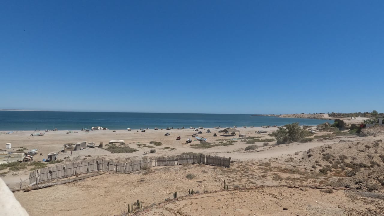 Baja California beach viewed from a sandy dune with a wooden fence in the foreground, scattered umbrellas and distant people on the sand, and blue ocean under a clear sky.