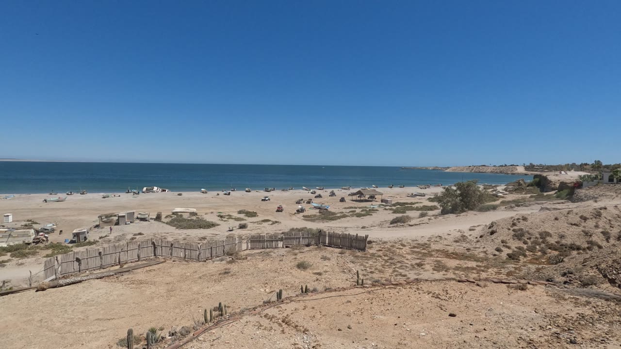 Wide beach and ocean beyond dry dunes with a wooden fence in the foreground.