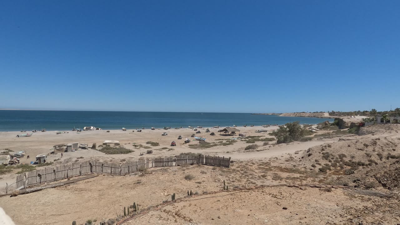 A dry Baja California dune and low wooden fencing frame a broad beach where small beachgoers and umbrellas sit between pale sand and the blue ocean.