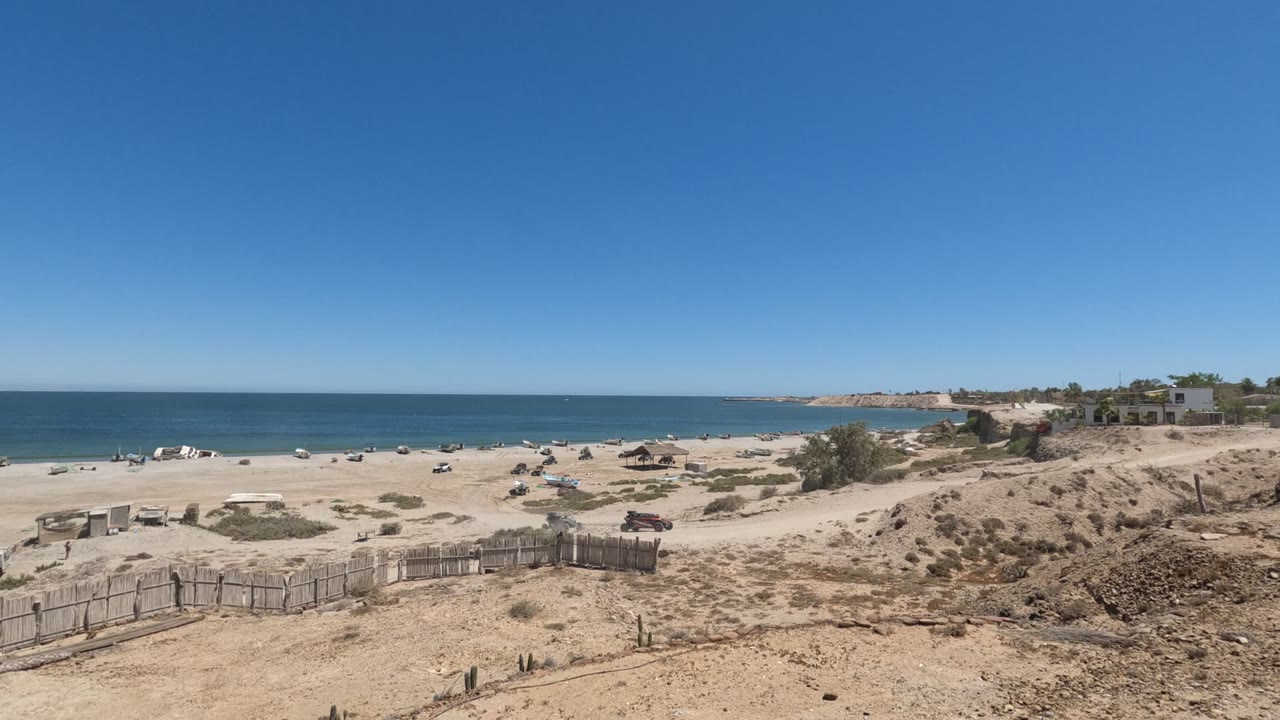 Wide Baja California, Mexico beach view from a sandy dune, with a wooden fence in the foreground, calm ocean beyond the shore, distant buildings, and no visible people at this moment.