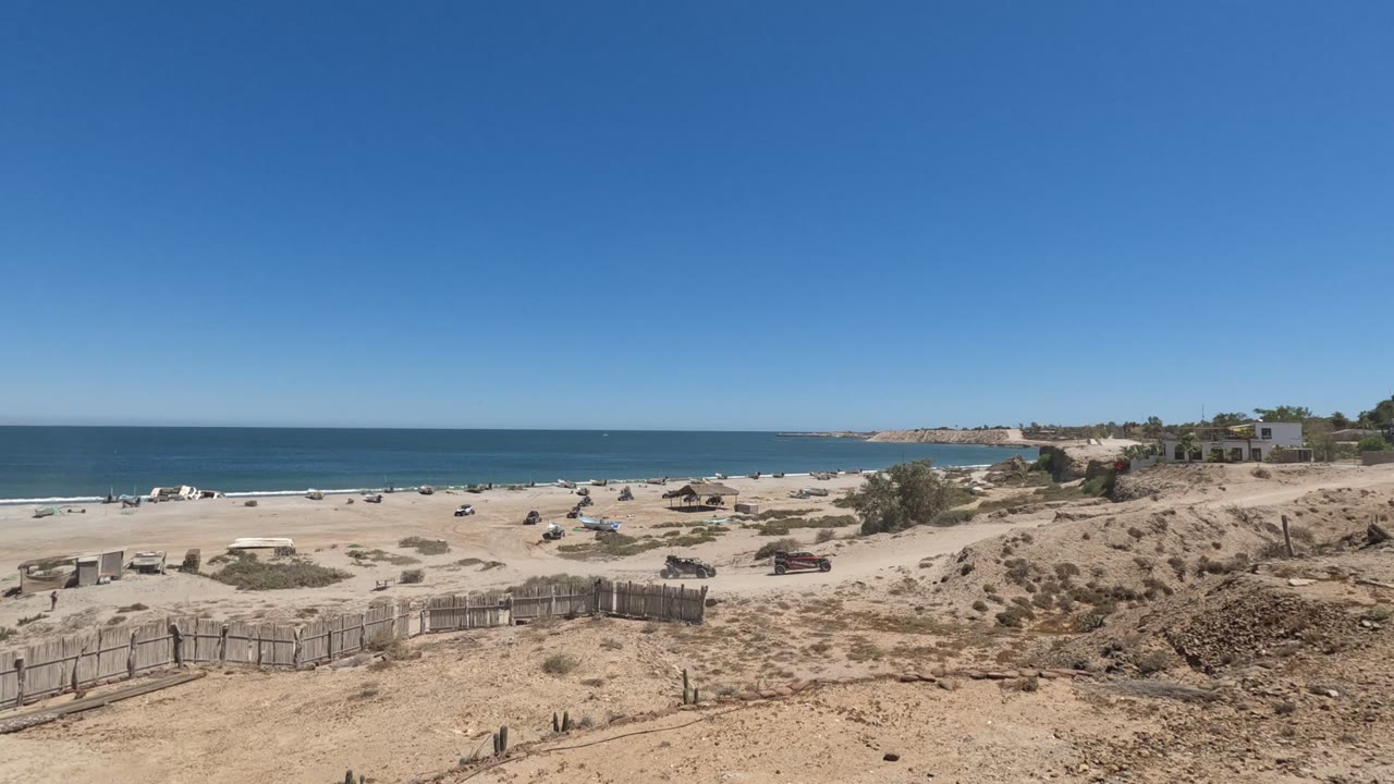 Wide Baja California, Mexico dune overlook showing the sandy foreground, weathered fence, small beach vehicles or structures, calm ocean water, distant shoreline, and no visible people.