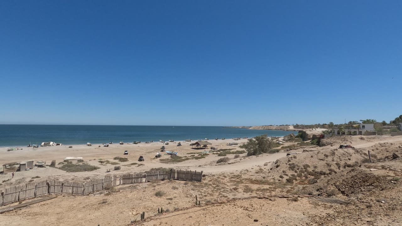 Wide Baja California, Mexico beach view at the start of the clip, with a wooden dune fence in the foreground, scattered people on the sand, and blue ocean beyond.