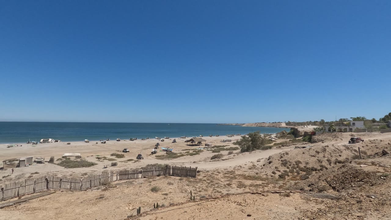 Sandy coast with low fence, dry scrub, beachgoers, and calm blue water.