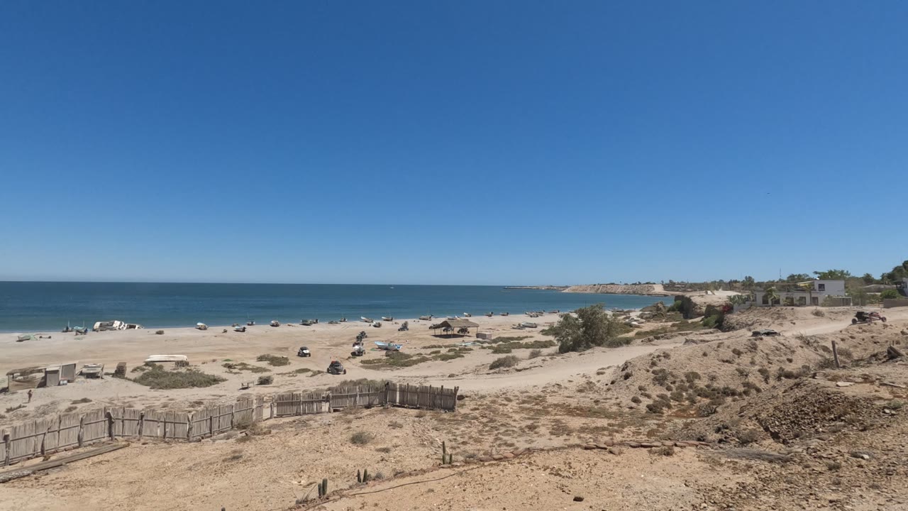 Wide Baja California, Mexico shoreline view with the dune fence low in frame, scattered beach items and people across the sand, and bright blue water under clear sky.