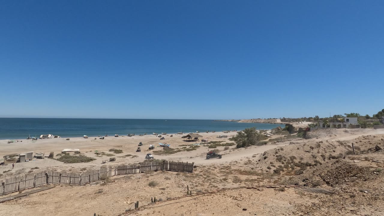 Opening frame of a Baja California sandy beach with a wooden fence in the foreground, vehicles near the shoreline, blue ocean beyond, and no visible people.