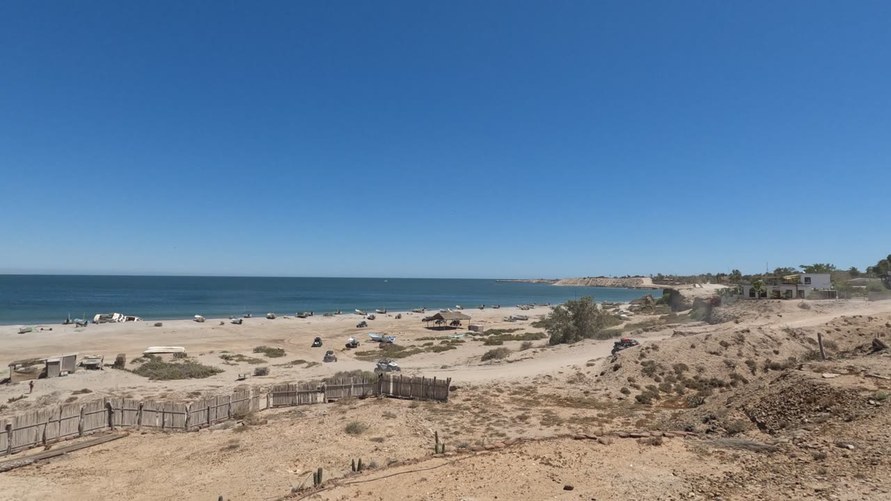 Wide Baja California coastal frame with dry dunes and rough fencing in front, scattered vehicles on the beach, blue water behind them, and no visible people.