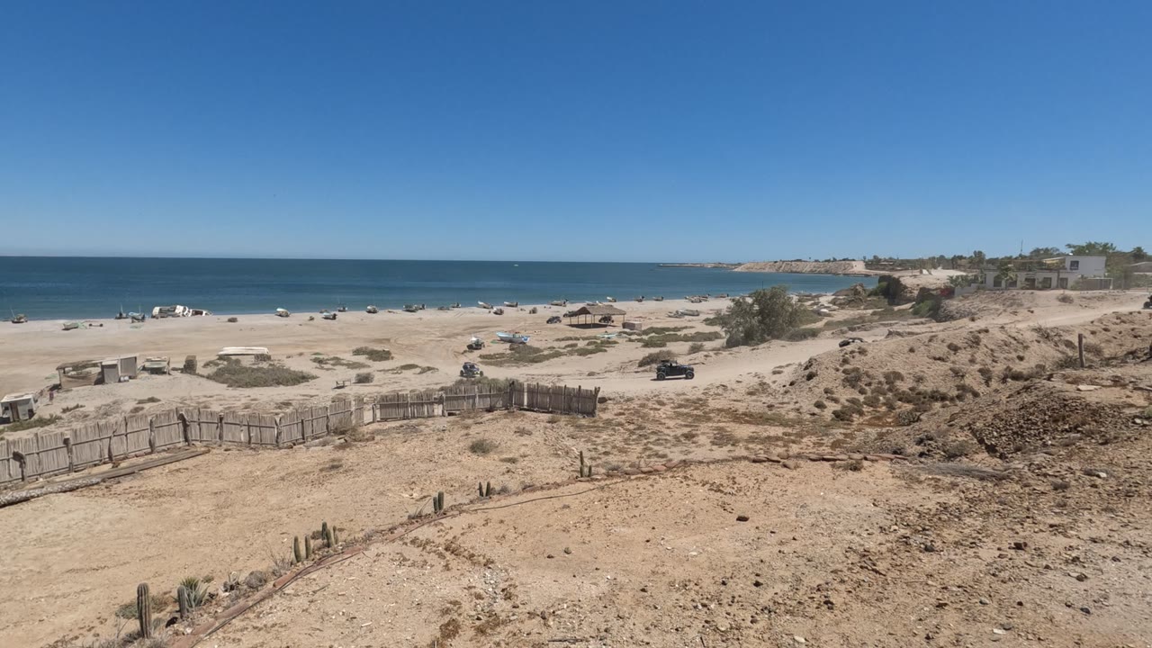 Wide view of a Baja California, Mexico beach with a rough fence in the foreground, ocean beyond the sand, a vehicle on the dirt path, and no visible people.