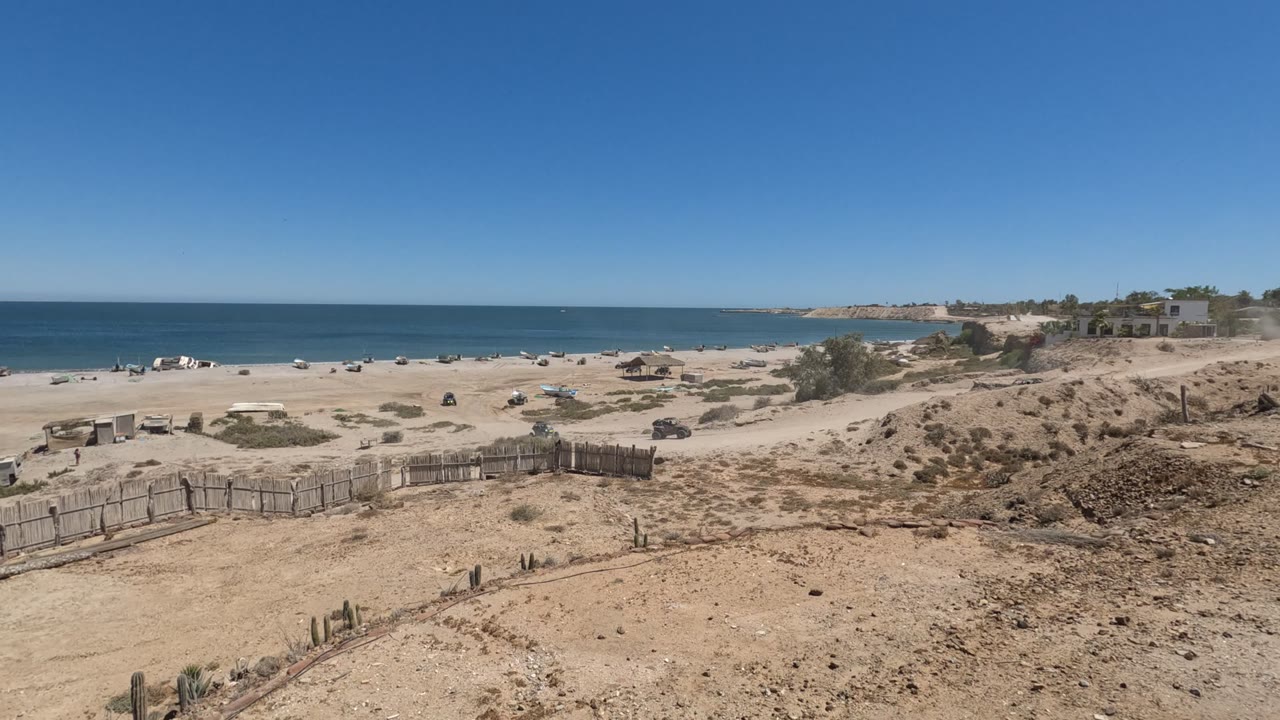 Wide Baja California coastline with the SUV near the right-side dunes, rustic fence panels in the foreground, beach shelters near the water, and no visible people.