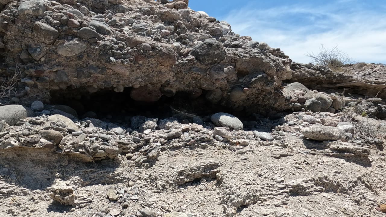 A rocky overhang in Baja California, Mexico casts shade over sandy, stone-covered ground beneath blue sky, with no visible people.