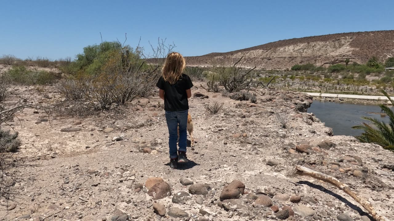 The dog is out of view as the girl walks ahead on the dry rocky path in Baja California, Mexico, with shrubs framing the trail and a narrow body of water to the right.